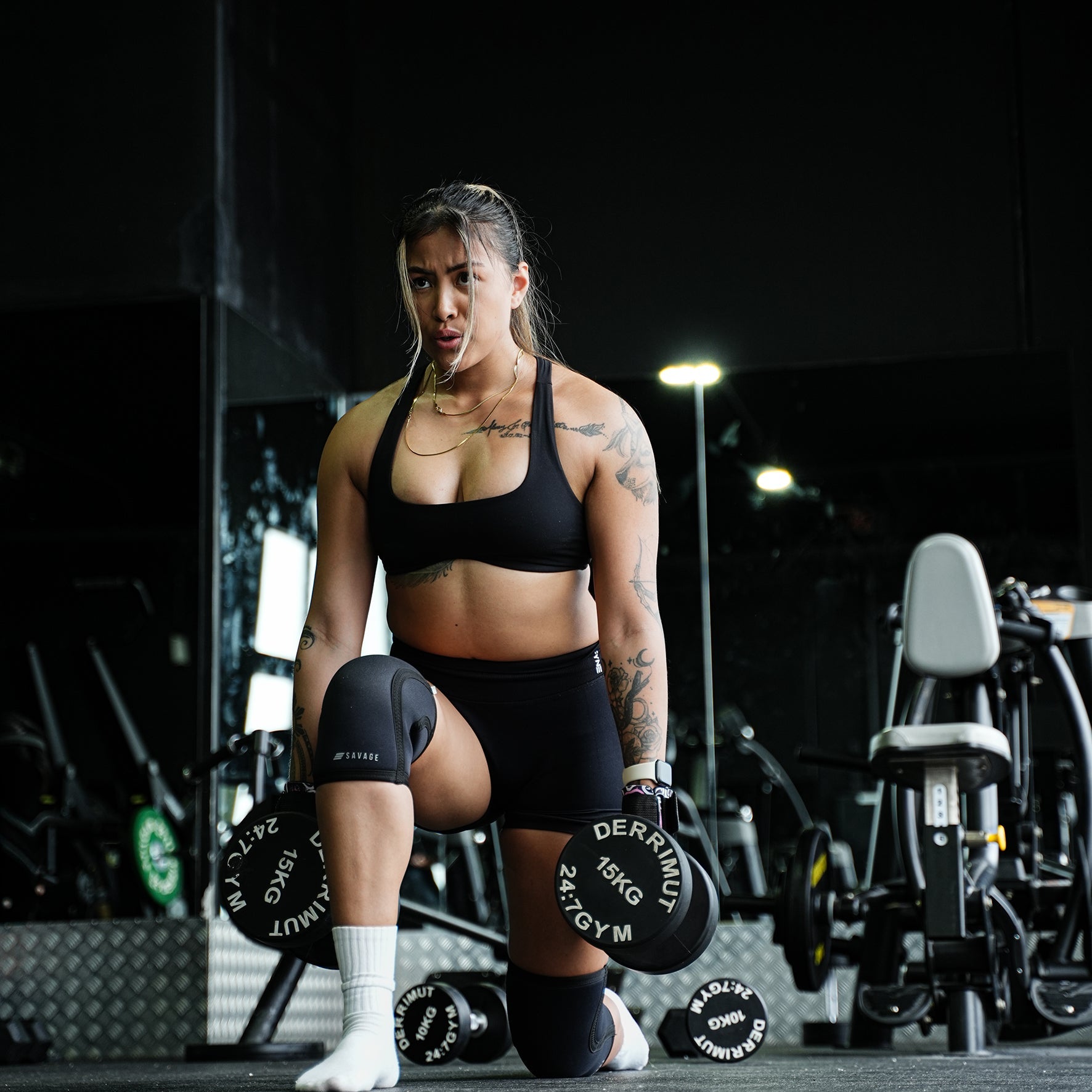 Woman lifting weights in a gym setting doing dumbbell lunges with lifting straps and wearing knee sleeves