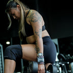 Woman lifting weights in a gym setting doing dumbbell lunges with lifting straps and wearing knee sleeves