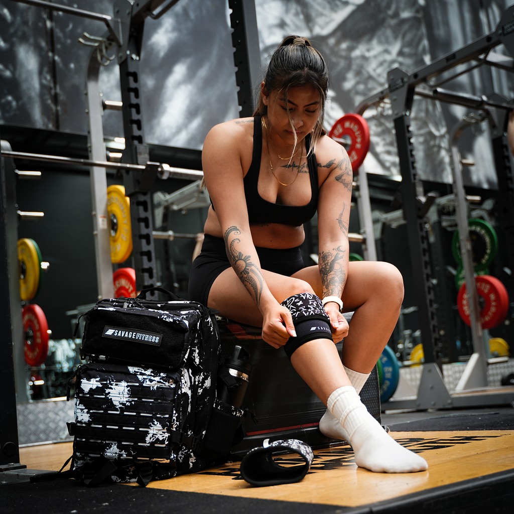Woman sitting on a weight bench in a gym setting putting on Savage Fitness Knee Sleeves