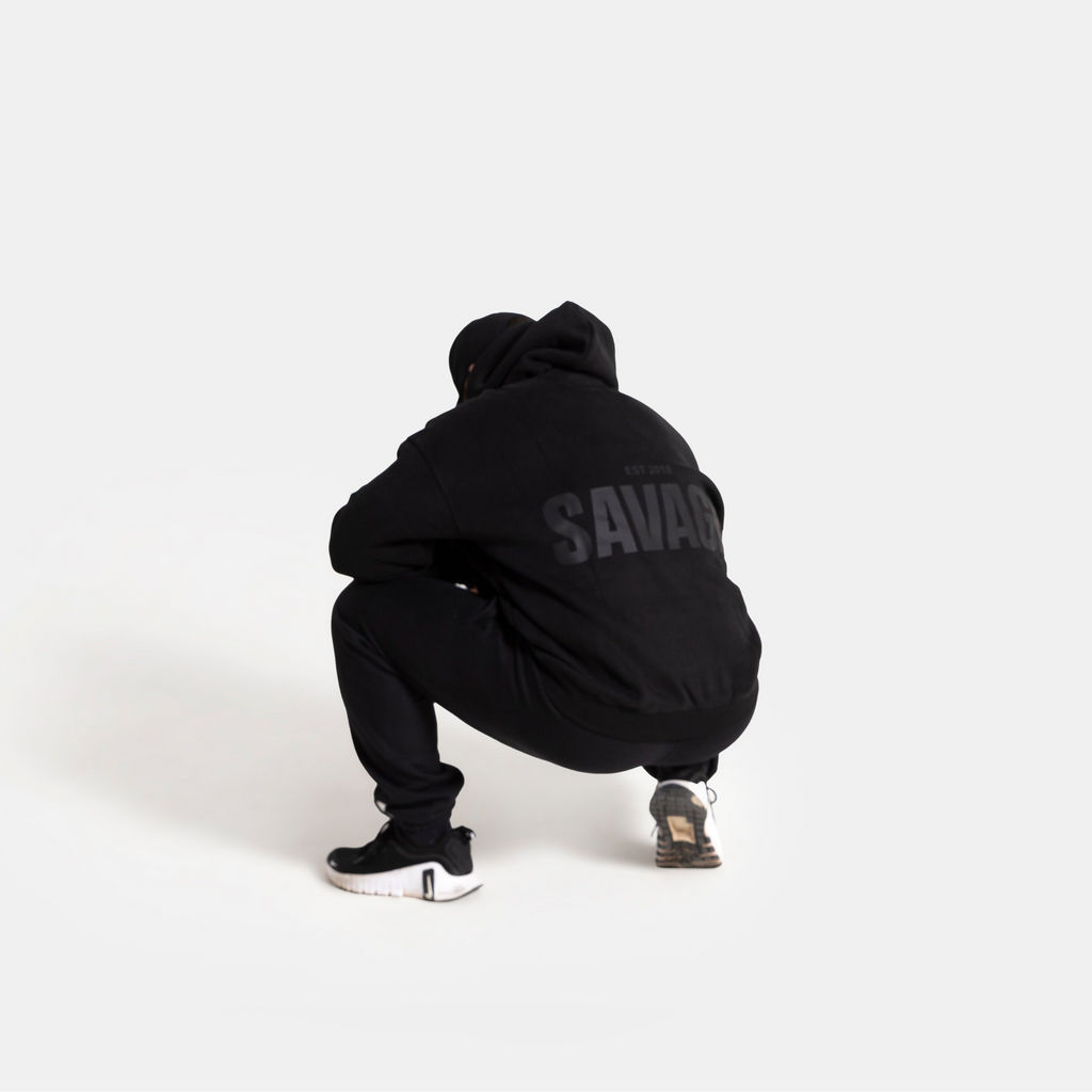 A person crouching from behind wearing a black hoodie with 'SAVAGE' printed in gray on the back, black sweatpants, and black sneakers, set against a solid light gray background.