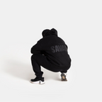 A person crouching from behind wearing a black hoodie with 'SAVAGE' printed in gray on the back, black sweatpants, and black sneakers, set against a solid light gray background.