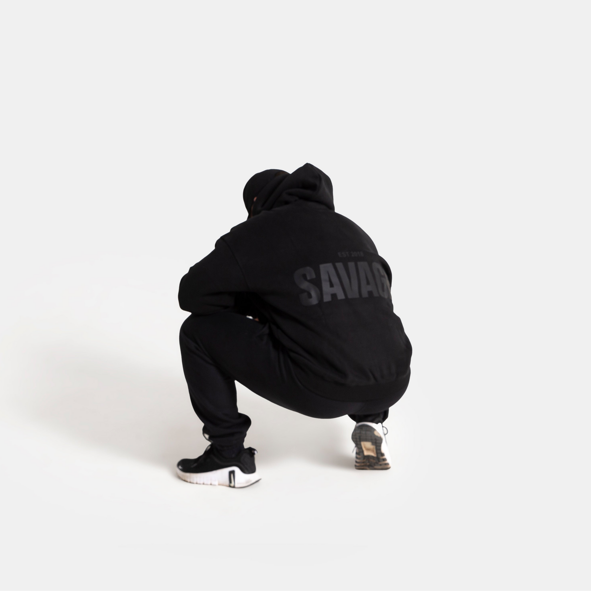 A person crouching from behind wearing a black hoodie with 'SAVAGE' printed in gray on the back, black sweatpants, and black sneakers, set against a solid light gray background.
