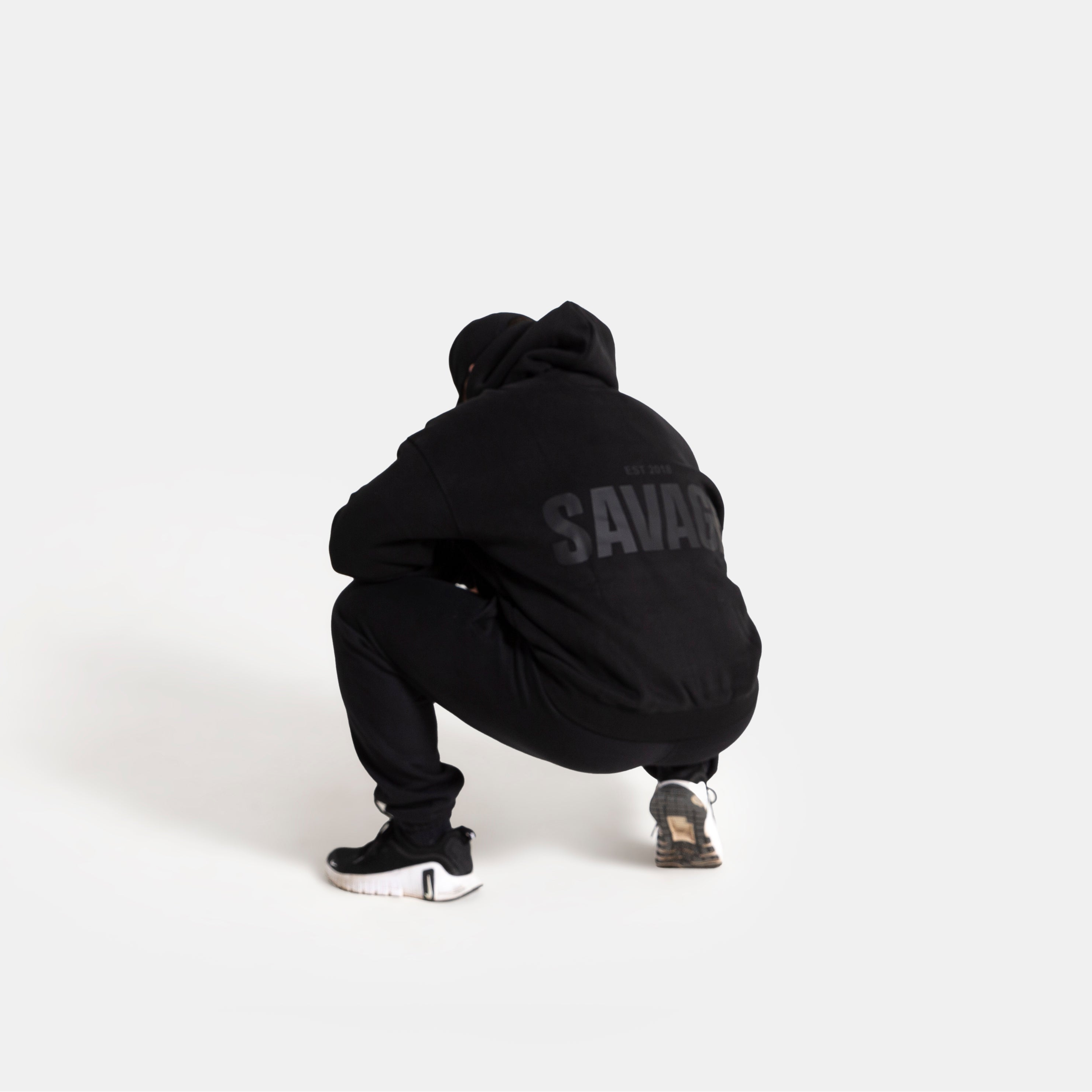 A person crouching from behind wearing a black hoodie with 'SAVAGE' printed in gray on the back, black sweatpants, and black sneakers, set against a solid light gray background.