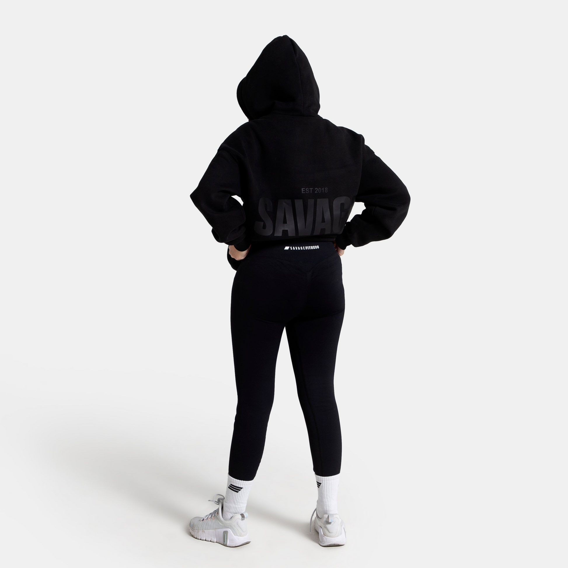 A model from behind wearing a black cropped hoodie with 'SAVAGE' printed in black on the back, paired with black leggings and white sneakers, against a plain white studio background.