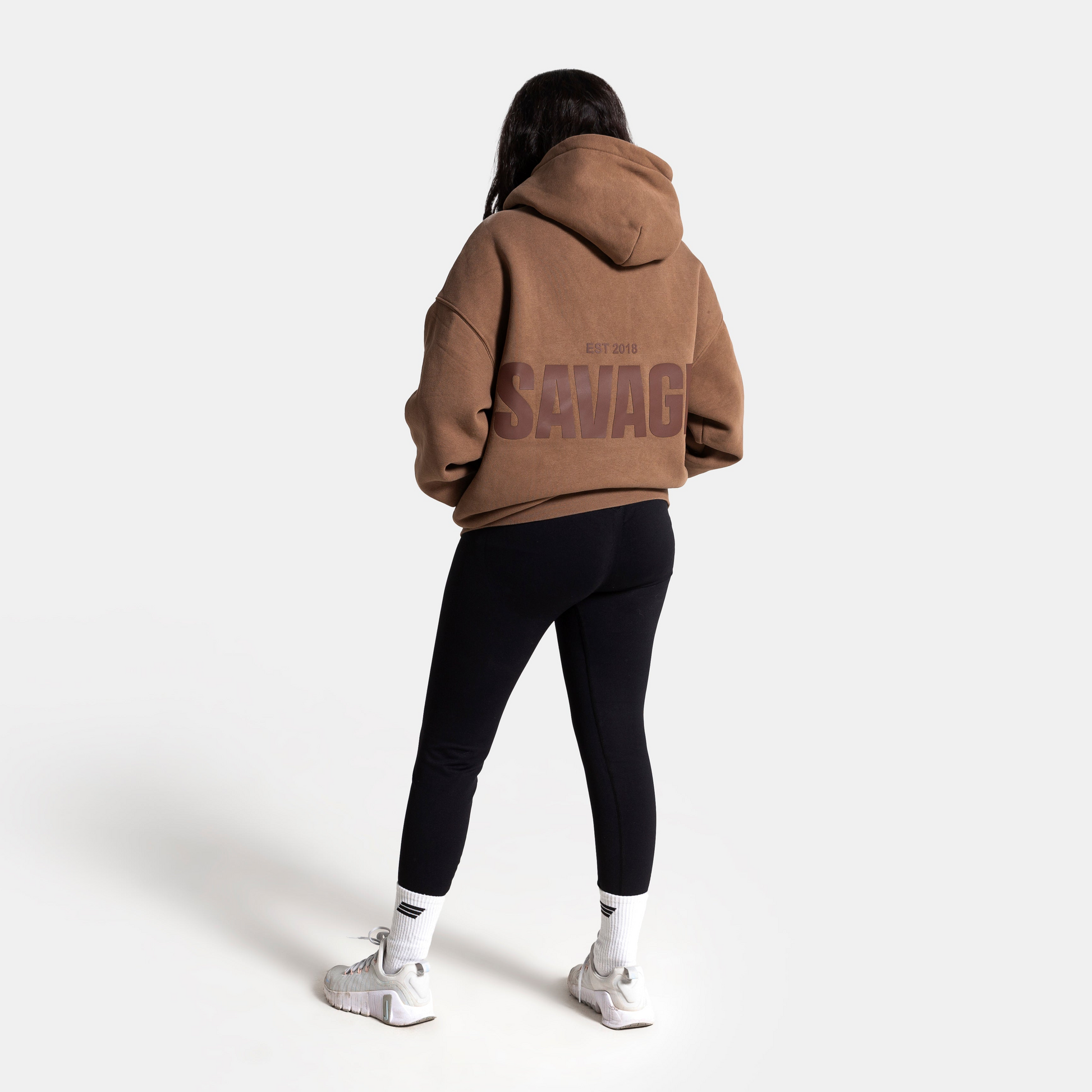 A model viewed from behind wearing a brown oversized hoodie with 'SEVAC' printed in dark letters on the back, paired with black leggings and white sneakers, against a plain white studio background.