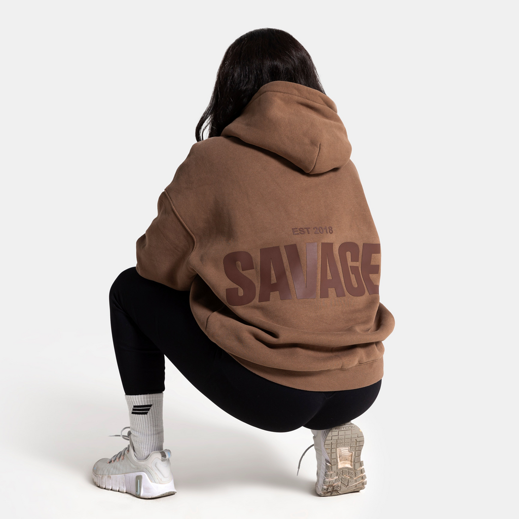 A person viewed from behind wearing an oversized brown hoodie with a large dark brown 'SAVAGE' graphic on the back, paired with black leggings and white sneakers, against a light gray studio background.