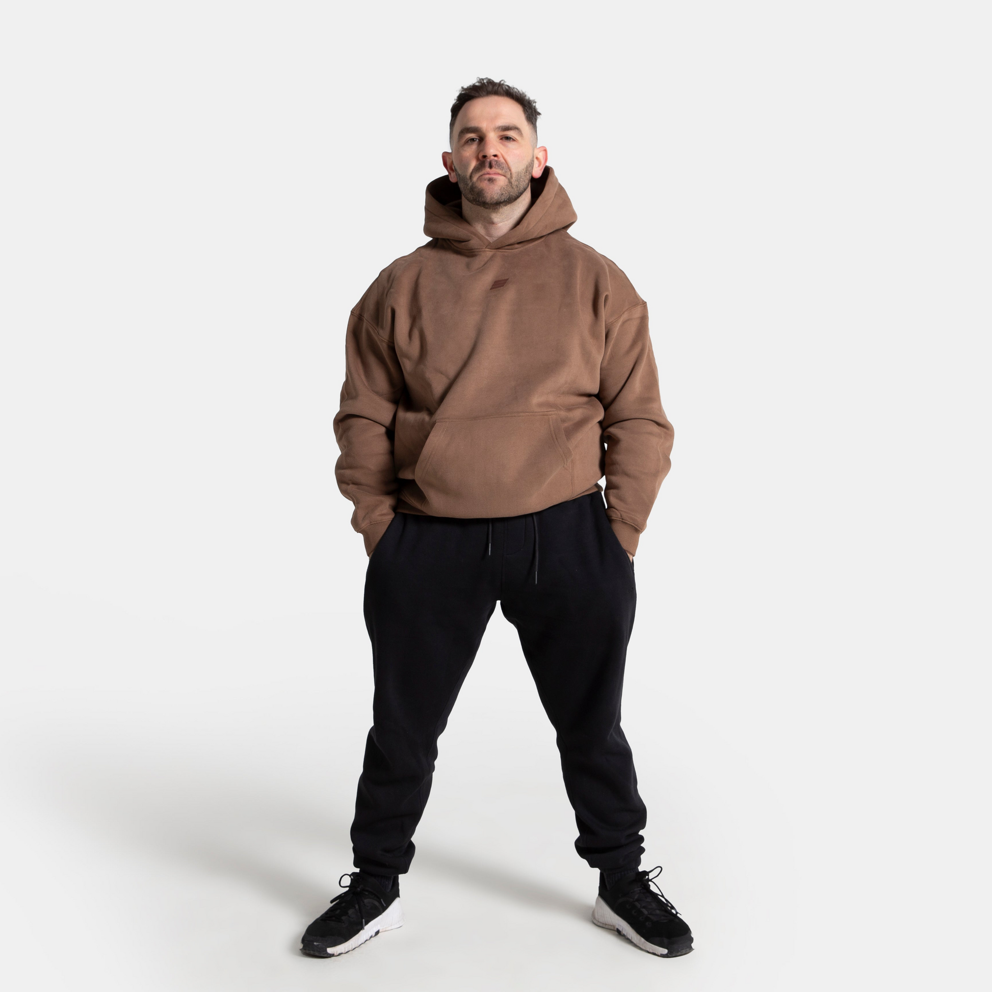 A man wearing a brown pullover hoodie and black sweatpants stands against a light gray studio background.
