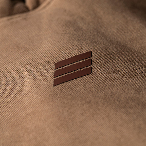 Close-up of a brown textured fabric garment with a small dark brown rectangular logo patch featuring Savage icon.