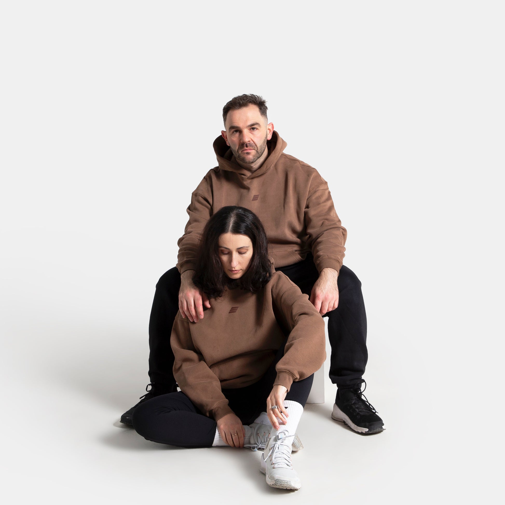A man and a woman in matching brown hoodies and black pants pose together on a seamless light gray studio background. The woman sits in front of the man, looking down at her white sneakers while tying them.