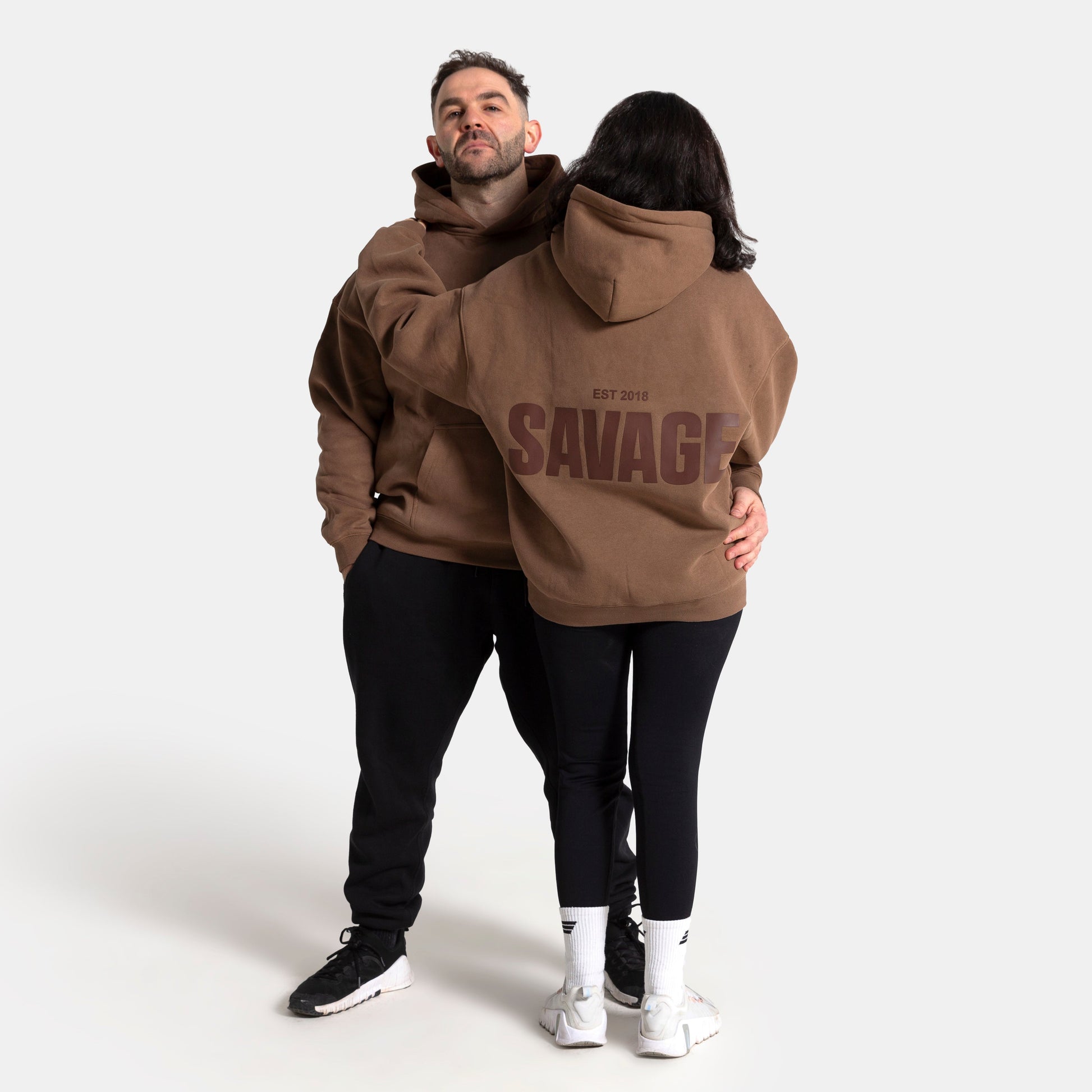 A man and a woman modeling matching brown hoodies with 'SAVAGE' printed in dark brown across the back, posed against a light gray studio background.