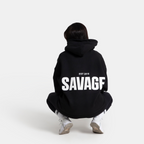 A person viewed from behind wearing a black hoodie with 'SAVAGF EST 2018' printed in bold white letters on the back, paired with black pants and white sneakers, set against a plain light gray background.