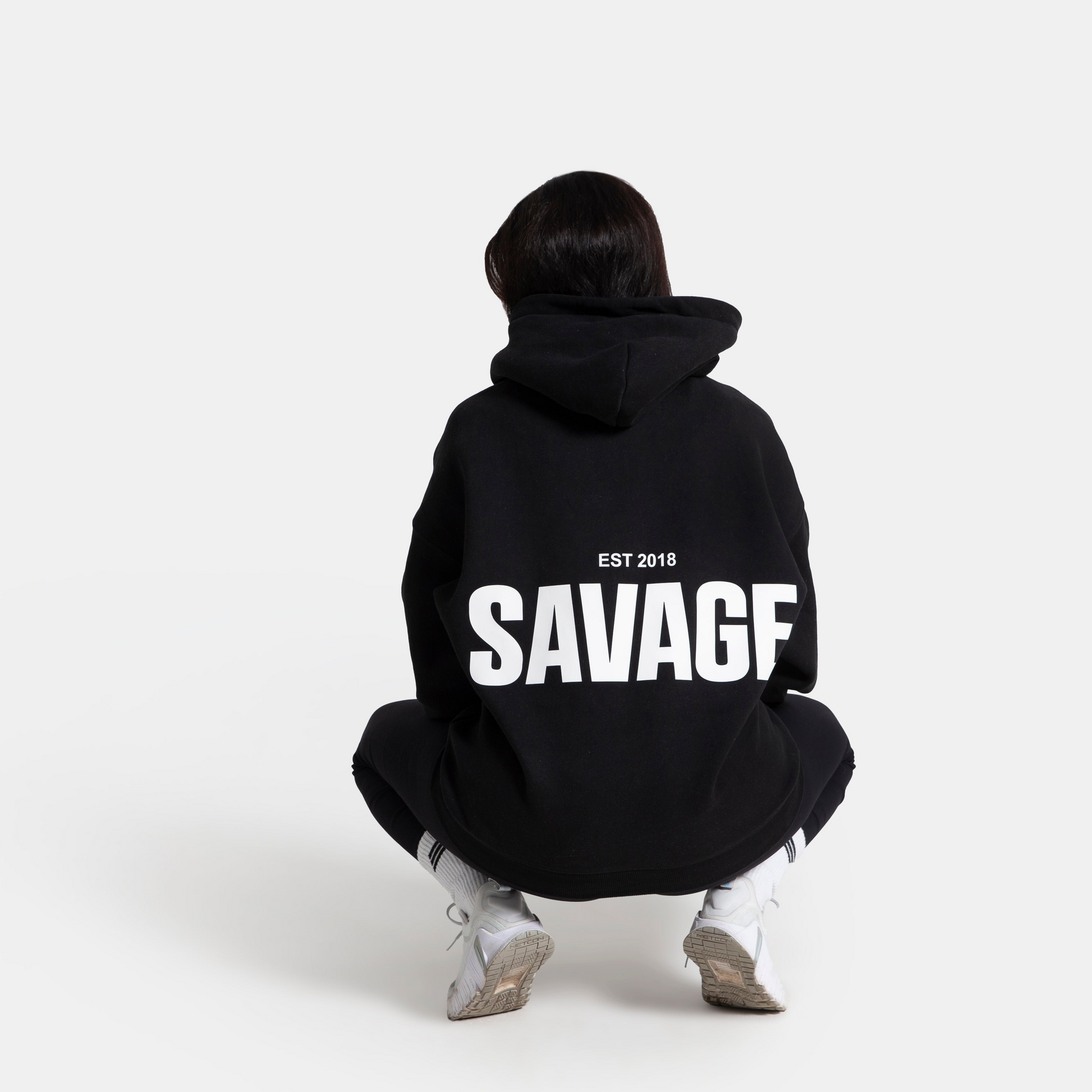 A person viewed from behind wearing a black hoodie with 'SAVAGF EST 2018' printed in bold white letters on the back, paired with black pants and white sneakers, set against a plain light gray background.