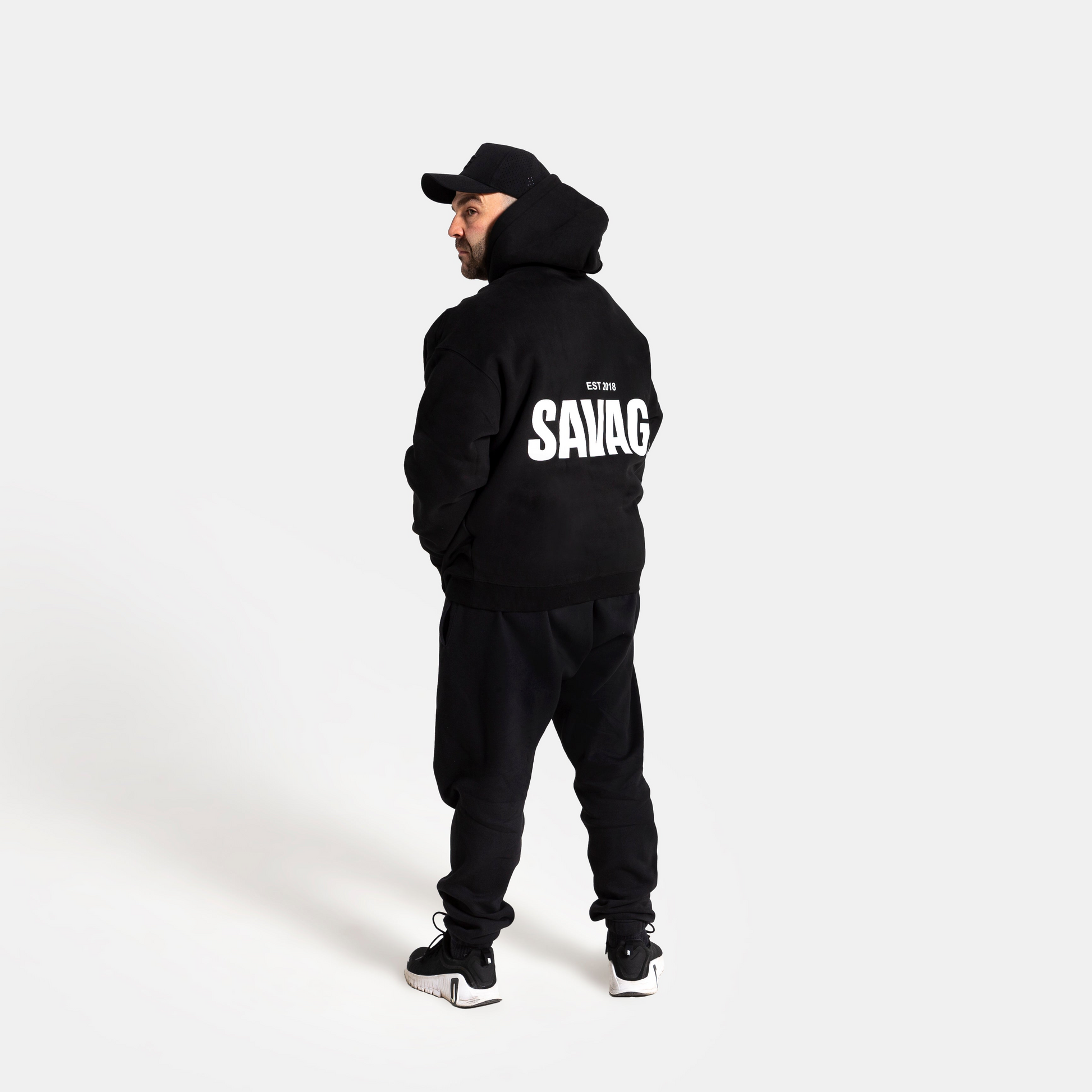 A man wearing a black hoodie with 'SAVAGE' printed in white on the back, black sweatpants, and a black cap, standing against a light gray background.
