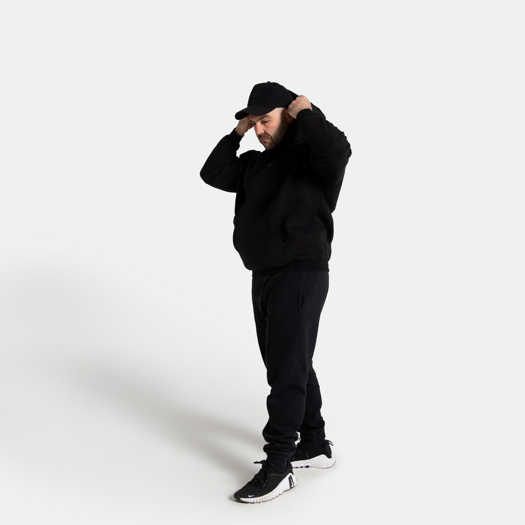 A person wearing a black hoodie, black sweatpants, a black cap, and black sneakers with white soles, standing against a light gray studio background.