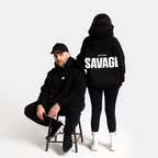 Two models in black streetwear clothing, one seated on a stool facing forward wearing a black cap and hoodie, and the other standing from the back wearing a black hoodie with 'SAVAGE' printed in bold white letters across the back.