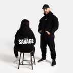 A man and a woman modeling black hoodies in a studio setting. The man stands wearing a black hoodie and matching black sweatpants, while the woman sits on a stool facing away from the camera, wearing a black hoodie with 'SAVAGE' printed in bold white letters on the back.