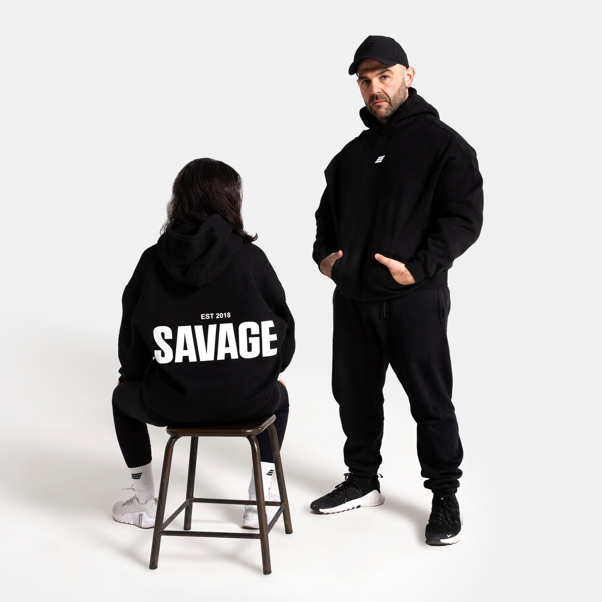 A man and a woman modeling black hoodies in a studio setting. The man stands wearing a black hoodie and matching black sweatpants, while the woman sits on a stool facing away from the camera, wearing a black hoodie with 'SAVAGE' printed in bold white letters on the back.