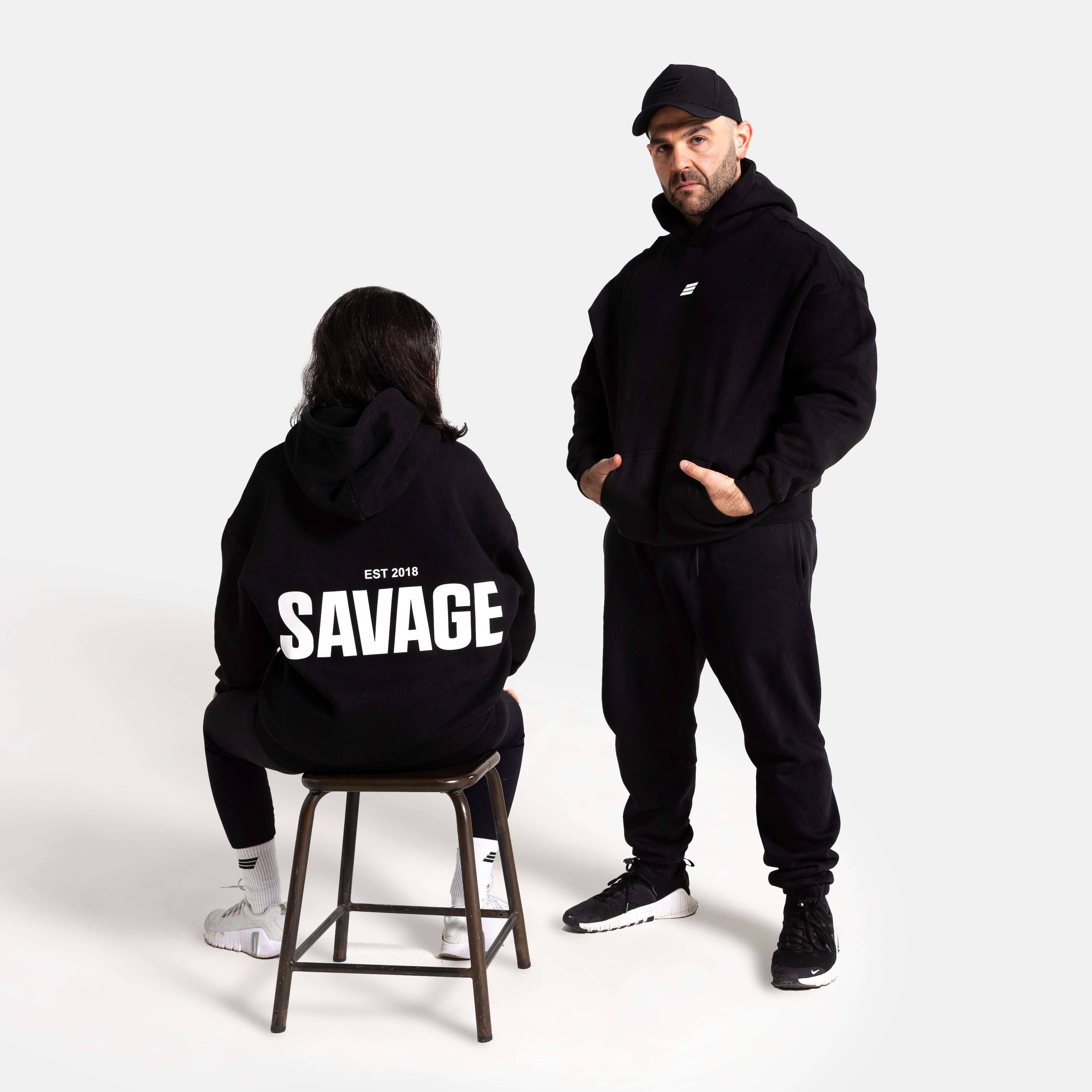 A man and a woman modeling black hoodies in a studio setting. The man stands wearing a black hoodie and matching black sweatpants, while the woman sits on a stool facing away from the camera, wearing a black hoodie with 'SAVAGE' printed in bold white letters on the back.