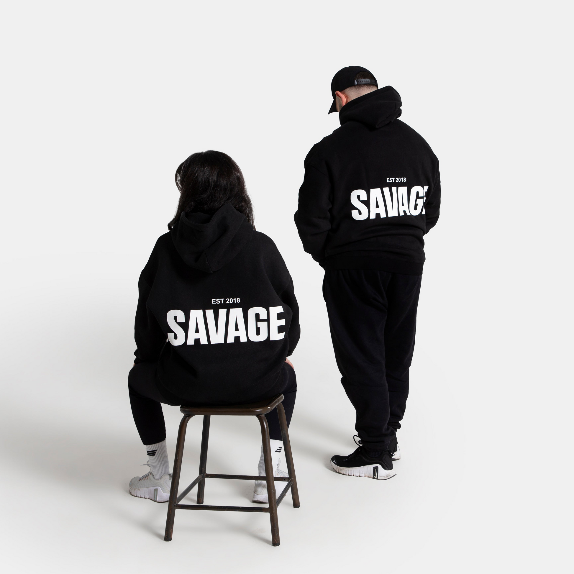 Two models shown from behind wearing matching black hoodies with a large white 'SAVAGE' graphic on the back, set against a plain light gray studio background.