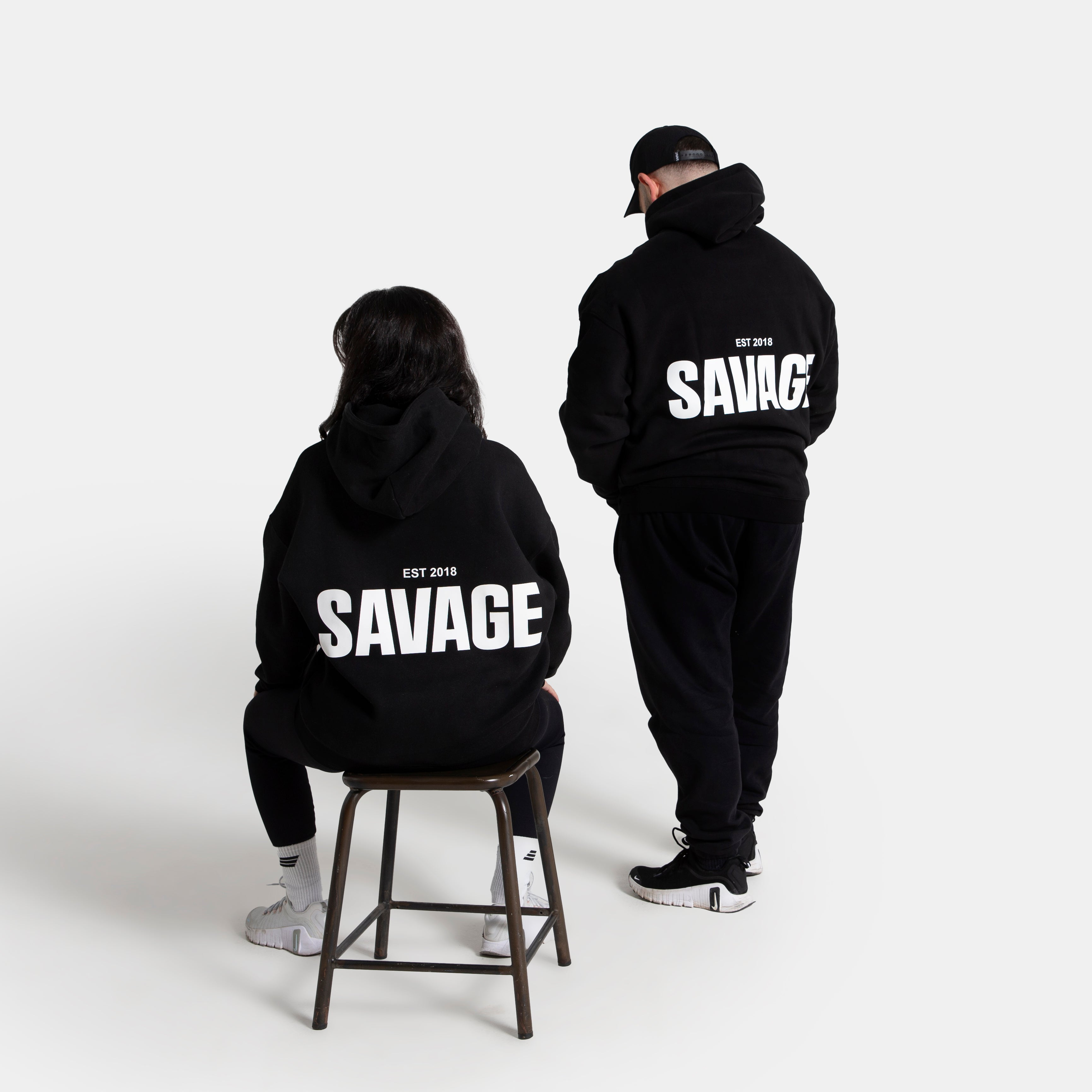 Two models shown from behind wearing matching black hoodies with a large white 'SAVAGE' graphic on the back, set against a plain light gray studio background.