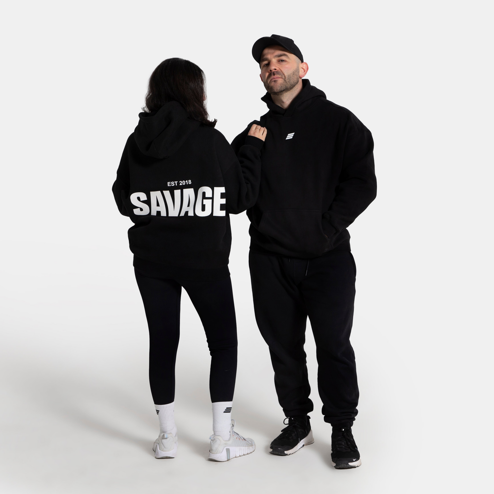 A man and a woman modeling matching black hoodies in a studio setting. The woman is shown from the back, wearing a black hoodie with 'SAVAGE EST 2019' printed in bold white letters, paired with black pants and white sneakers.