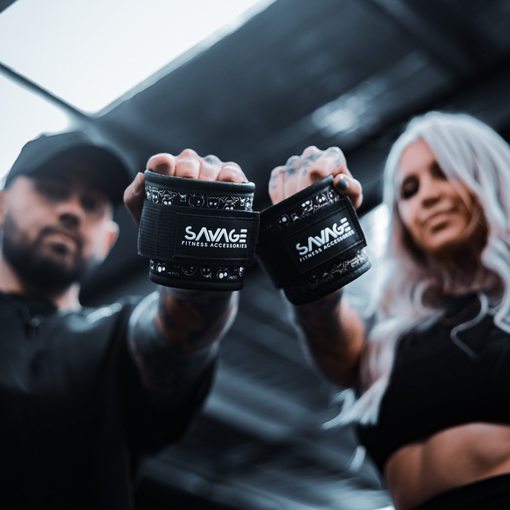 Two people holding black ankle wraps with 'Savage Fitness Accessories' branding in a gym setting.