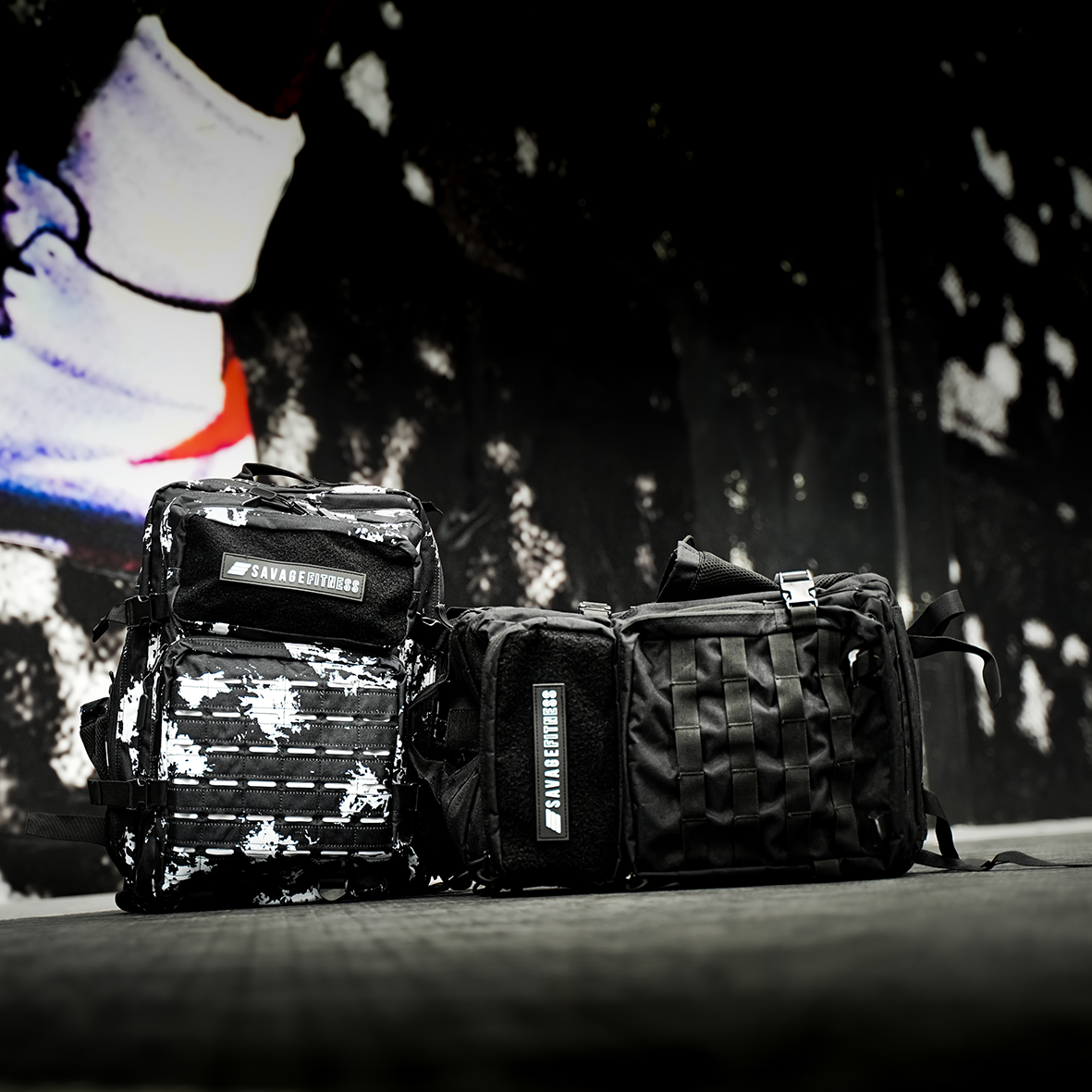 Two Savage Fitness
 tactical bags with visible branding on a dark background with a blurred image of a person wearing white sneakers.