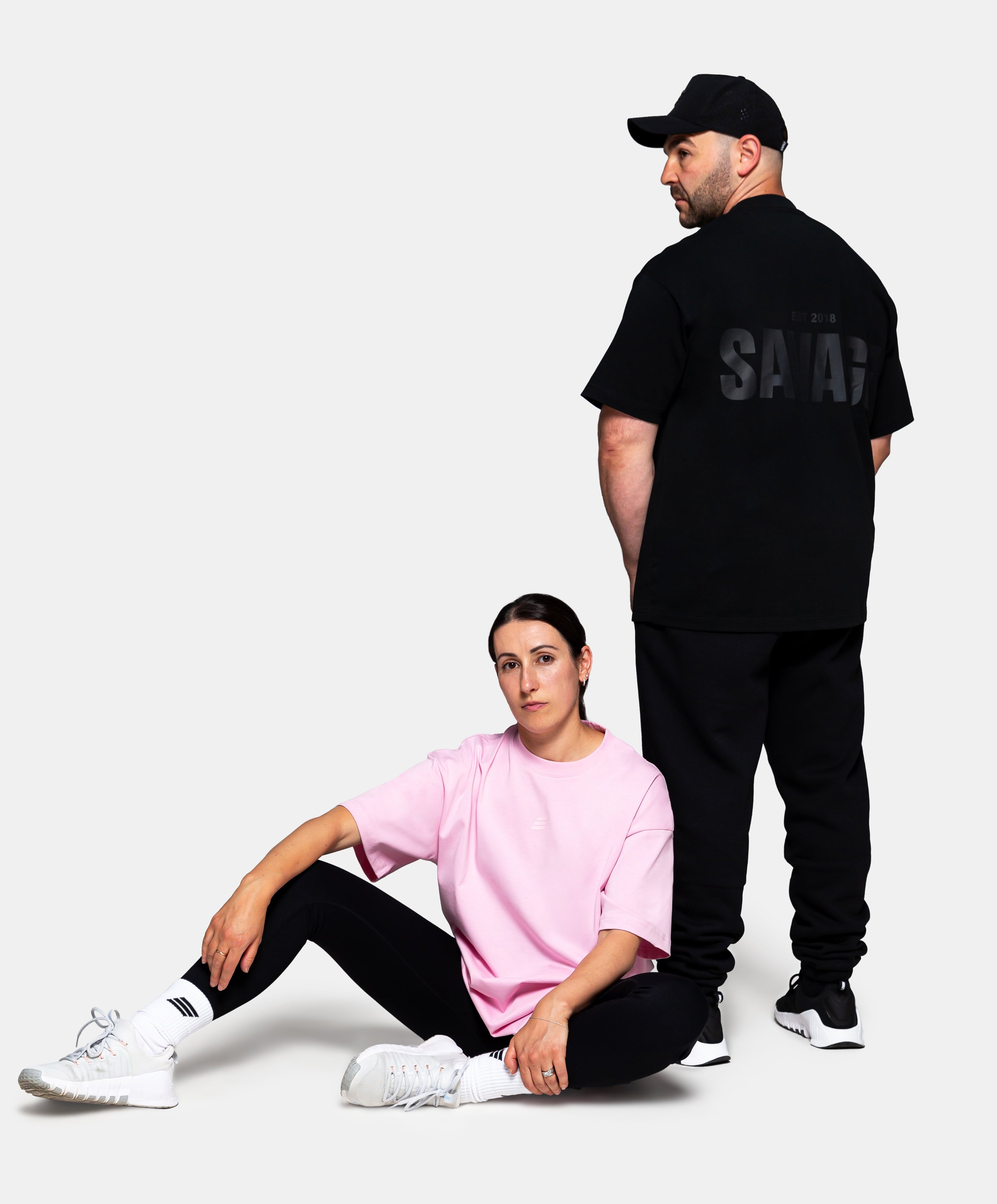 Savage Fitness oversized tee in pink on female model seated front view and black oversized tee on male model showing back print in studio setting